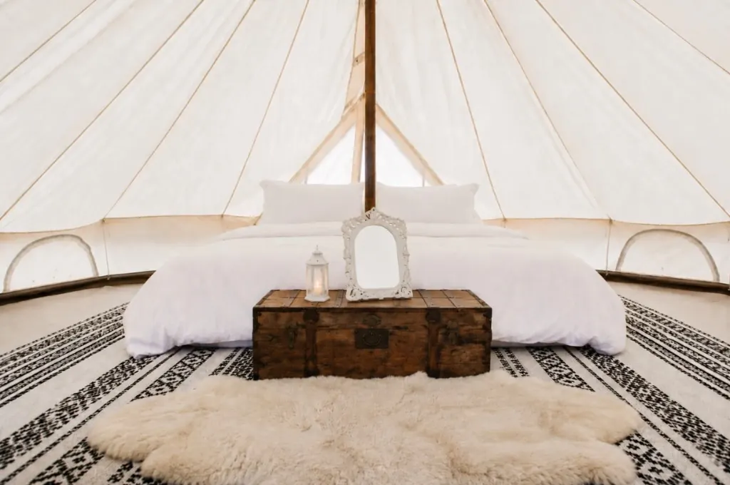 Interior of a luxury glamping bell tent with white linens, a rustic trunk with lantern and mirror, and a sheepskin rug